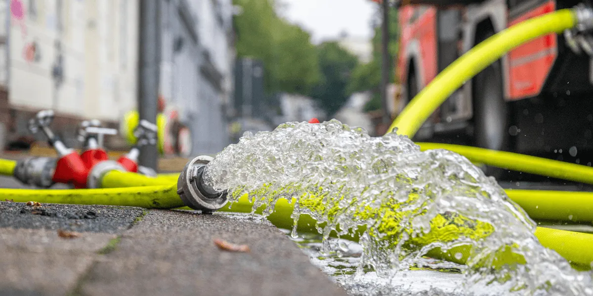 LöRüRl: Nahaufnahme eines leuchtend gelben Feuerwehrschlauchs, aus dessen Metallkupplung auf einer Straße kräftig Wasser austritt; im unscharfen Hintergrund steht ein Feuerwehrfahrzeug.