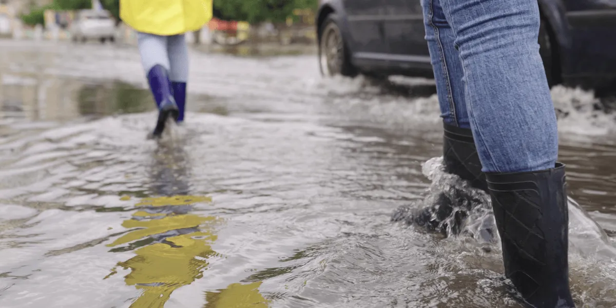Drainagebeton: Personen gehen mit Gummistiefeln durch eine überflutete Straße, während ein Auto durch das Wasser fährt. Eine Person trägt einen gelben Regenmantel.