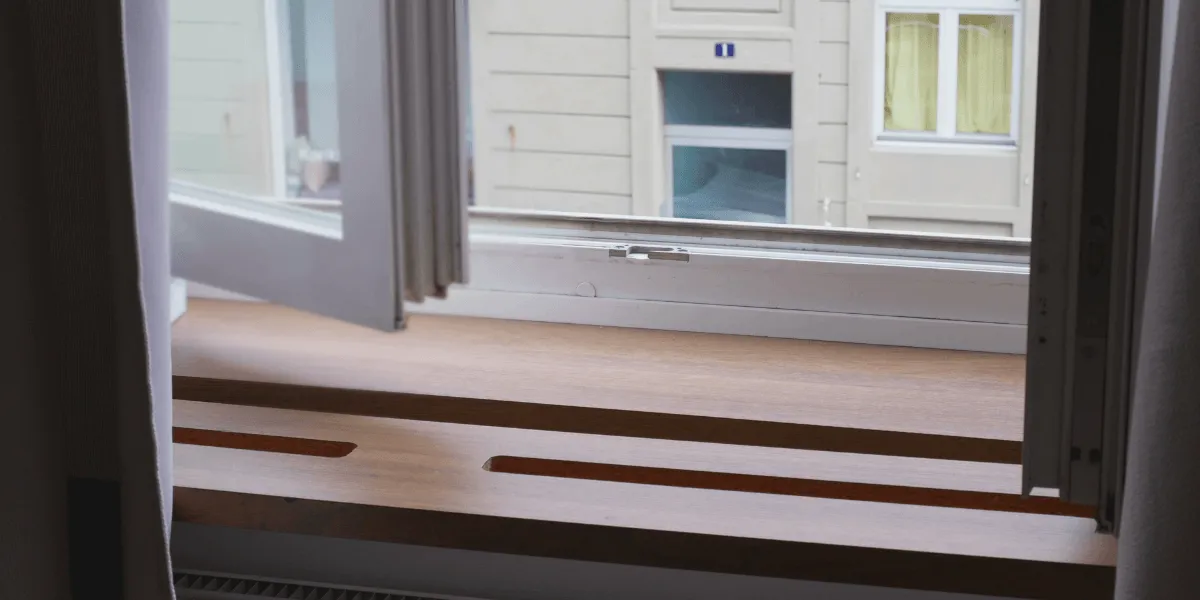Wohnraumlüftung: Blick von innen auf eine breite Holzfensterbank vor einem geöffneten Fenster; gegenüber ist die Fassade eines Nachbargebäudes zu sehen.