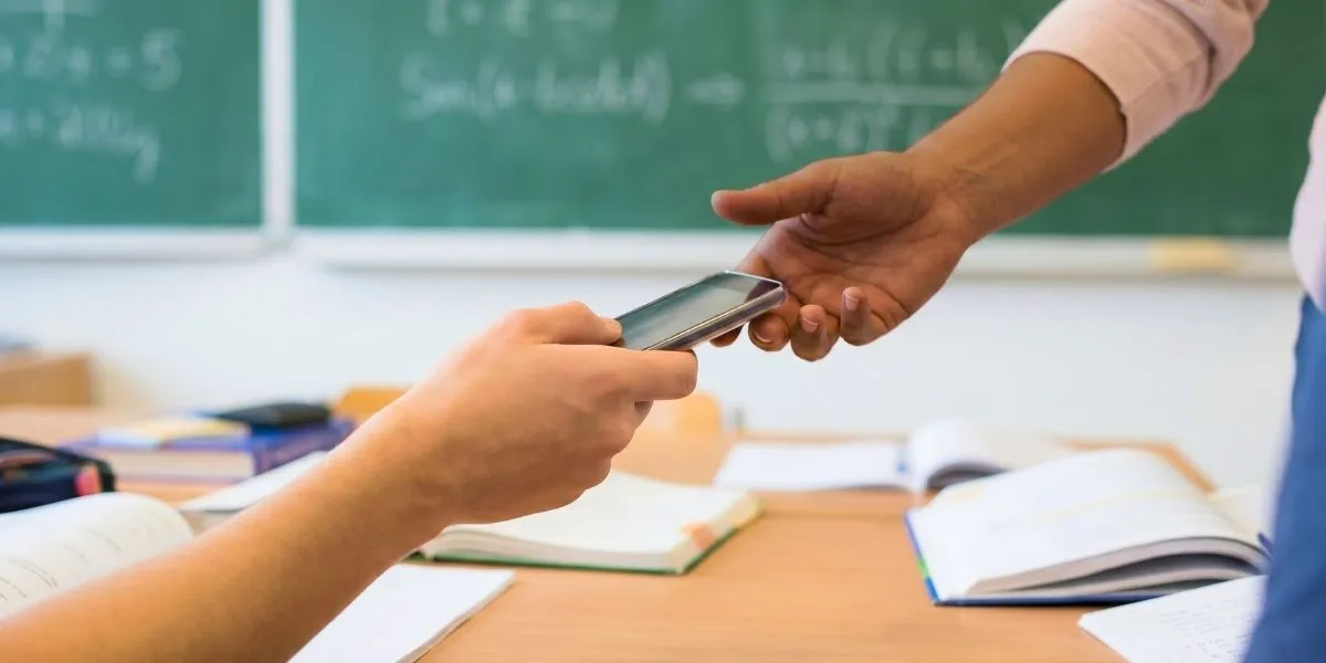 Nahaufnahme der Hand einer Schülerin, die ihrer Lehrerin ihr Handy übergibt wegen des Handyverbots an Schulen