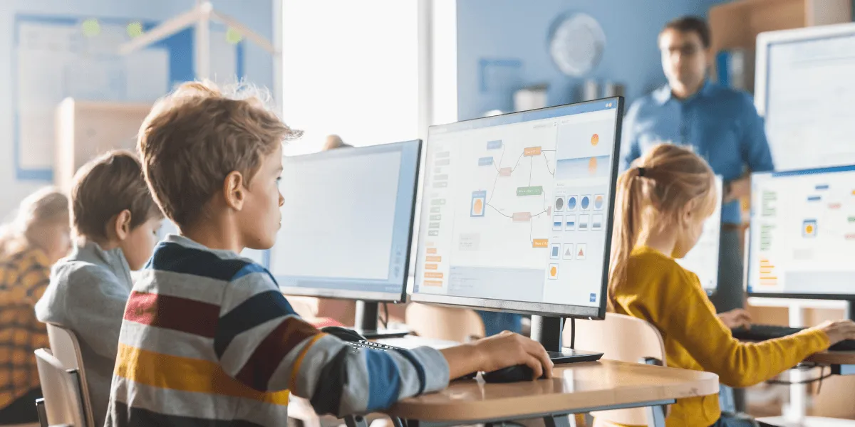 Unterrichtsphasen: Schülerinnen und Schüler arbeiten in einem Computerraum an großen Monitoren; im Hintergrund steht eine Lehrkraft. Auf einem Bildschirm ist ein digitales Flussdiagramm zu sehen.