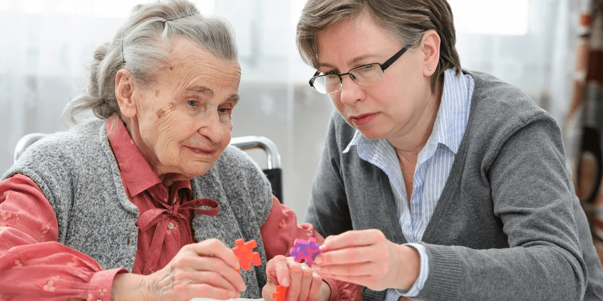 Expertenstandard Demenz: Eine ältere Frau mit grauem Haar und eine jüngere Betreuerin mit Brille sitzen an einem Tisch und lösen gemeinsam ein buntes Kunststoff-Puzzle als Beschäftigungs- und Gedächtnistraining.