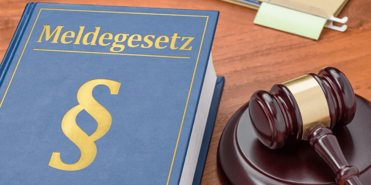 Erweiterte Melderegisterauskunft: Blaues Gesetzbuch mit der Aufschrift „Meldegesetz“ und goldenem Paragrafenzeichen neben einem Richterhammer auf einem Holztisch.