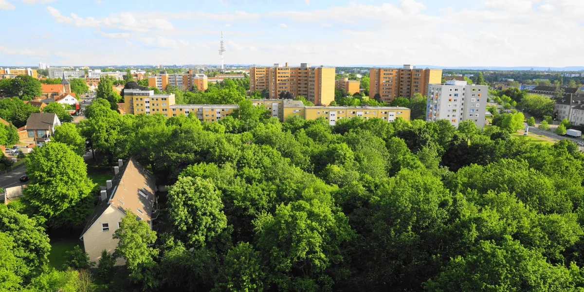 Stadtbegrünung: Luftaufnahme eines begrünten Stadtviertels mit dichtem Baumbestand, Wohnhäusern und mehrgeschossigen Gebäuden unter blauem Himmel.