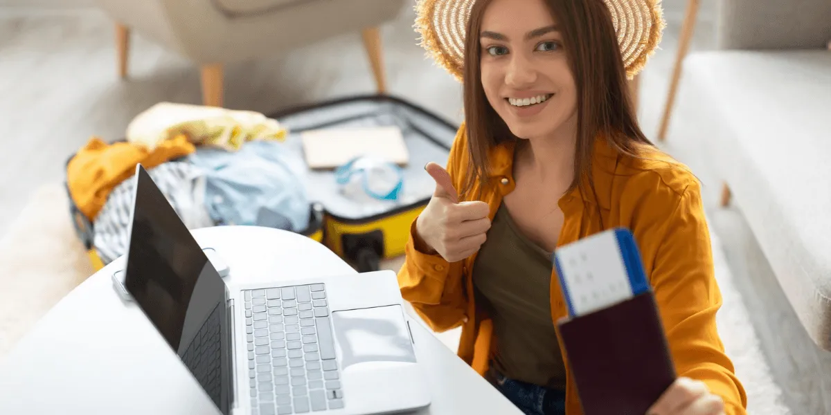 Lächelnde junge Frau in legerer Kleidung, die einen Reisepass mit Flugtickets in der Hand hält, während sie zu Hause am Laptop arbeitet. Sie freut sich auf die bevorstehende Reise und ihre Urlaubspläne, die Szene spielt in einem modernen Wohnzimmer.