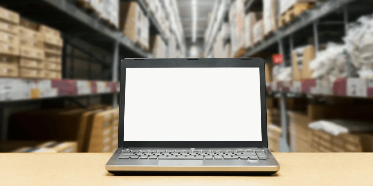 Endverbleibserklärung: Laptop mit leerem Bildschirm auf einem Tisch vor hohen Lagerregalen mit Kartons und Paletten.