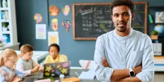 Classroom Management: Grundschullehrer im Klassenzimmer schaut in Kamera, hinter ihm arbeiten Kinder an einer Gruppenarbeit.an einer Gruppenarbeit.