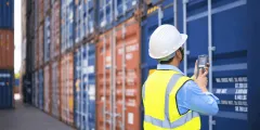 EORI Nummer: Ein Mitarbeiter in Warnweste und Schutzhelm scannt mit einem mobilen Gerät einen blauen Frachtcontainer auf einem Containerlagerplatz.