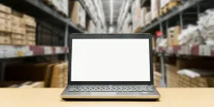 Endverbleibserklärung: Laptop mit leerem Bildschirm auf einem Tisch vor hohen Lagerregalen mit Kartons und Paletten.