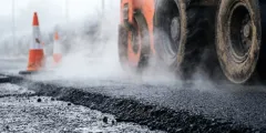Ersatzbaustoffverordnung: Eine Straßenwalze fährt über frisch aufgetragenen Asphalt; Dampf steigt von der heißen Fahrbahn auf, im Hintergrund stehen mehrere orange-weiße Verkehrskegel.