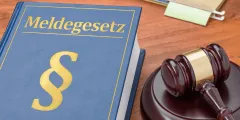 Erweiterte Melderegisterauskunft: Blaues Gesetzbuch mit der Aufschrift „Meldegesetz“ und goldenem Paragrafenzeichen neben einem Richterhammer auf einem Holztisch.