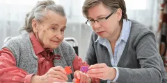 Expertenstandard Demenz: Eine ältere Frau mit grauem Haar und eine jüngere Betreuerin mit Brille sitzen an einem Tisch und lösen gemeinsam ein buntes Kunststoff-Puzzle als Beschäftigungs- und Gedächtnistraining.