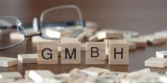 GmbH gründen: Holzbuchstaben mit der Aufschrift „GMBH“ stehen auf einem Tisch zwischen weiteren Buchstabensteinen. Im Hintergrund liegt eine Brille.