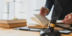 Nahaufnahme des Schreibtisches und der Hände einer Anwältin, die in einem Gesetzbuch blättert, vor ihr auf dem Tisch ein Richterhamme, neben ihr eine Waage