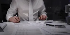 IDW S1: Person in weißem Hemd und Krawatte prüft und unterschreibt Dokumente an einem Schreibtisch neben Taschenrechner, Lupe und Laptop.