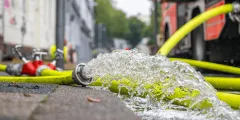 LöRüRl: Nahaufnahme eines leuchtend gelben Feuerwehrschlauchs, aus dessen Metallkupplung auf einer Straße kräftig Wasser austritt; im unscharfen Hintergrund steht ein Feuerwehrfahrzeug.