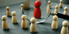 Talent Management: Rote Holzfigur im Fokus einer Lupe, umgeben von hellen Holzfiguren auf dunkler Fläche.
