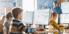Unterrichtsphasen: Schülerinnen und Schüler arbeiten in einem Computerraum an großen Monitoren; im Hintergrund steht eine Lehrkraft. Auf einem Bildschirm ist ein digitales Flussdiagramm zu sehen.