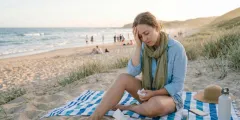 30-jährige Frau sitzt an Nordseestrand auf einer Decke im Urlaub, ist aber erkältet und hat Kopfschmerzen, hält ein Taschentuch in einer Hand