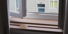 Wohnraumlüftung: Blick von innen auf eine breite Holzfensterbank vor einem geöffneten Fenster; gegenüber ist die Fassade eines Nachbargebäudes zu sehen.