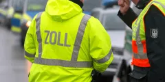 Zollfreigrenze: Zollbeamte in Warnjacken bei einer Straßenkontrolle, ein Fahrzeug im Hintergrund.